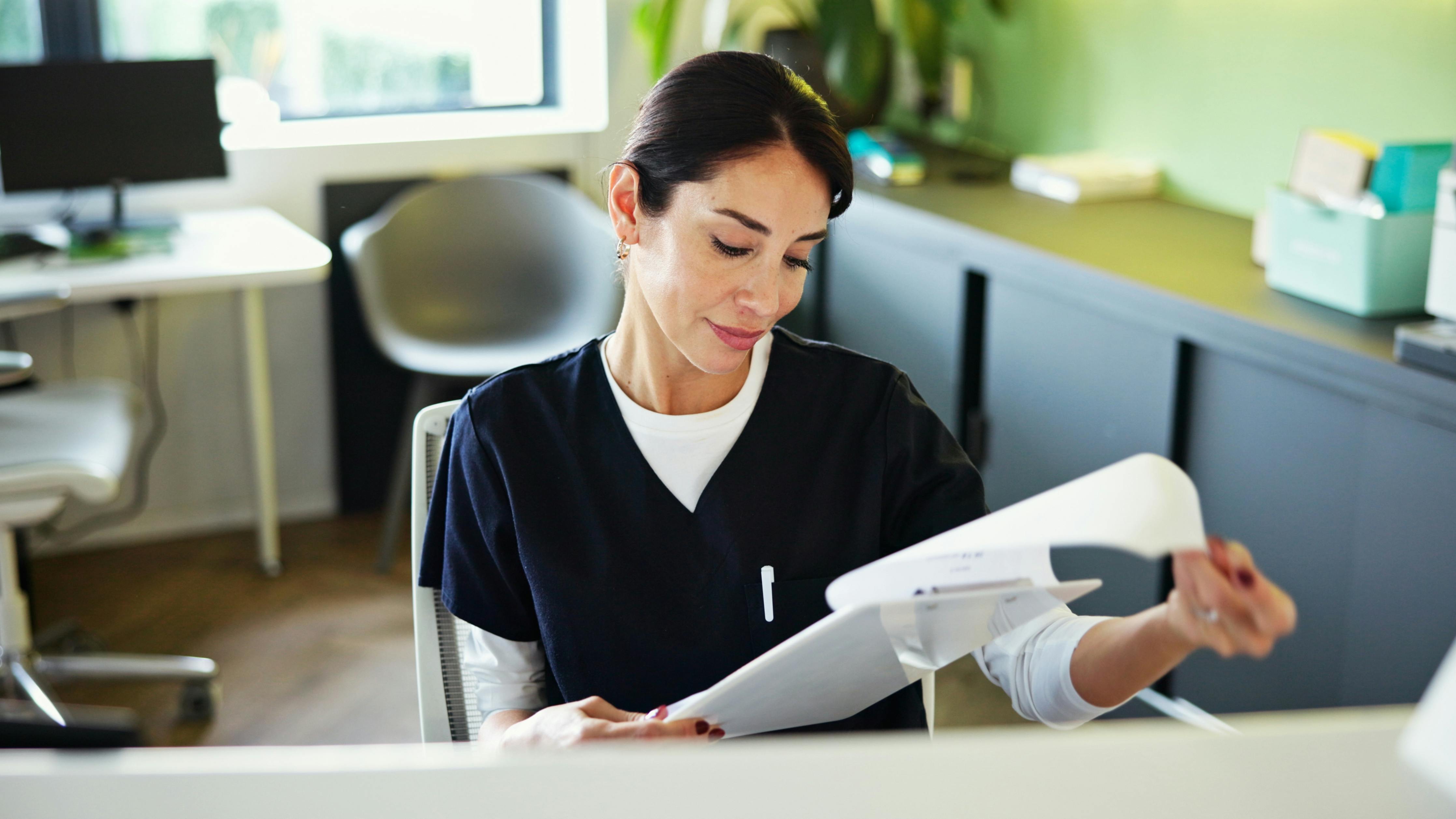 Woman in scrubs sits at front desk and reviews paperwork on a clipboard