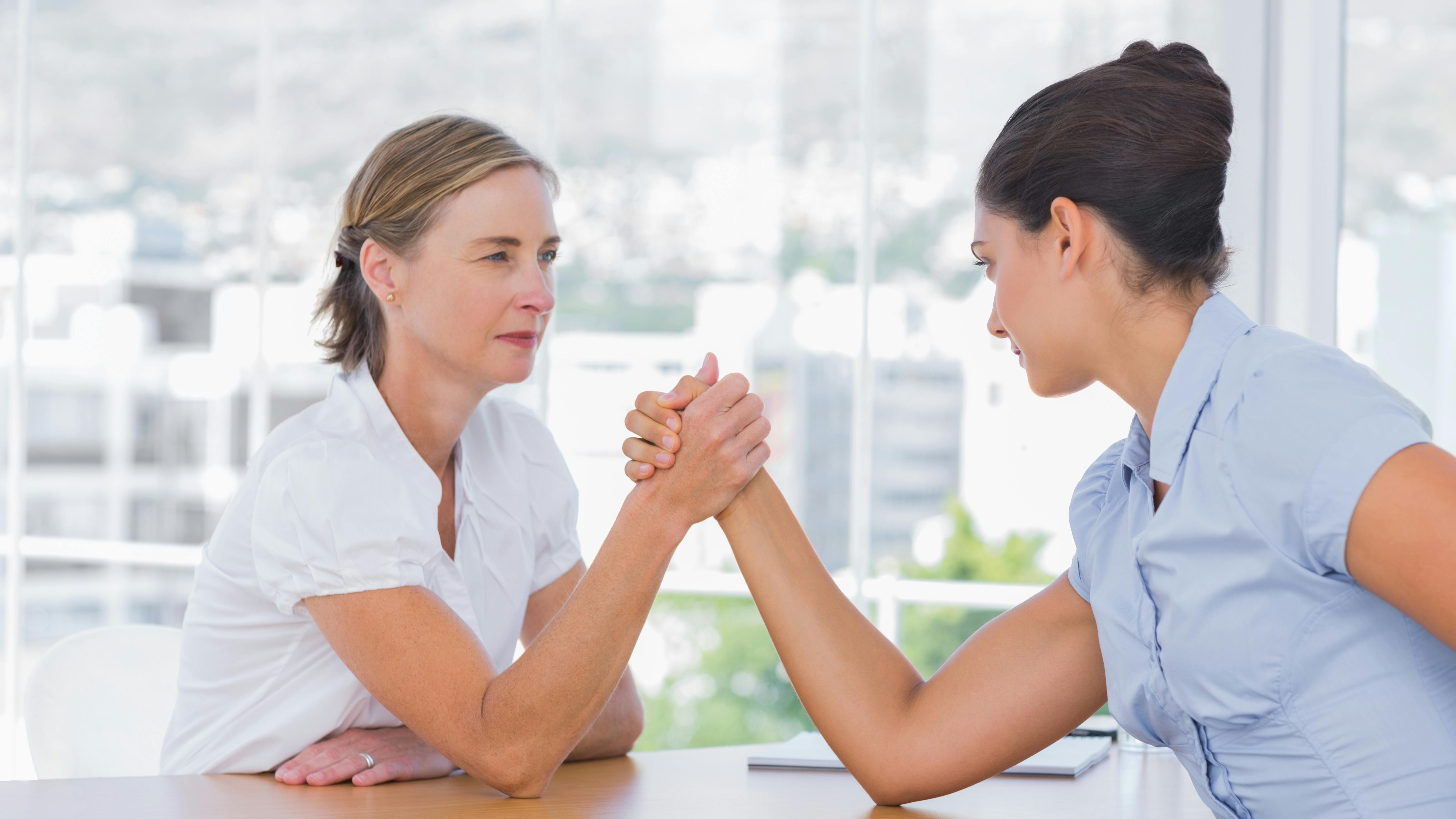 Two businesswomen sitting at a table arm-wrestling