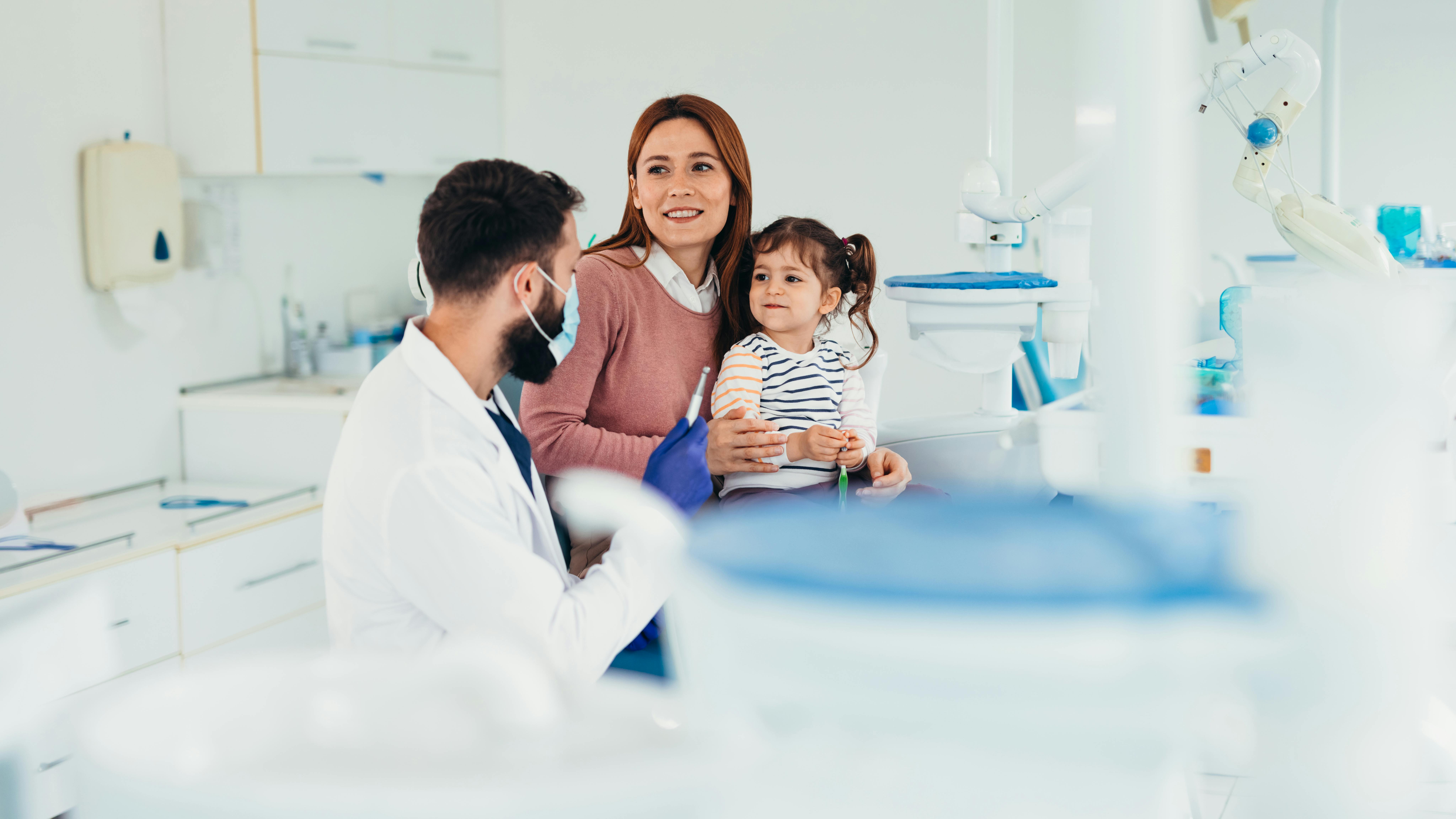 Dentist wearing face mask talking with mother and daughter during appointment in dental office, explaining treatment and giving advice for oral hygiene